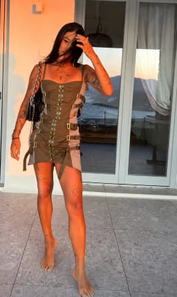 The woman stands with her weight shifted onto one leg, her hips angled and torso upright, one arm raised so her hand rests against the side of her head with fingers threaded through her hair while the other hangs loosely at her side. She wears a tightly fitted mini dress in olive and taupe panels, its bodice shaped by vertical rows of gold eyelets and lacing that pull inward at the waist, with additional straps and buckles wrapping the torso and hips. The abbreviated hem leaves her thighs and legs bare, the skin smooth and evenly toned, the muscles of her calves and thighs visibly defined under the warm light. Her bare feet rest on the stone floor with the left angled forward and toes relaxed while the right heel lifts slightly to reveal the smooth sweep from heel to ball, warm-toned skin carrying a light natural sheen across the tops and inner arches along with faint tension lines on the raised arch and subtle creases at the base of the toes. Glossy green and bright blue alternating polish covers each toenail and finishes with a crisp white checkered overlay that contrasts against her skin, and add a sexy, girly, and flirtatious edge to her look which is absolutely stimulation, arousing, and appealing in appearance, while a delicate script tattoo marks the outer left ankle and a small dark emblem sits on the inner right ankle just above the bone. Small script and illustrative tattoos mark the length of her legs, adding detail to the otherwise uninterrupted line from hip to ankle. Her legs and feet call out as being absolutely receptive for eroticism and sex in a highly pleasing manner.